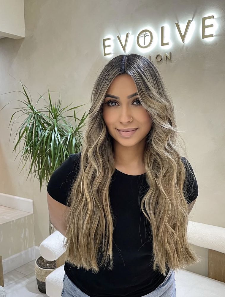 Waist-length mushroom blonde balayage with dark roots, styled in loose center-parted waves at Evolve Salon